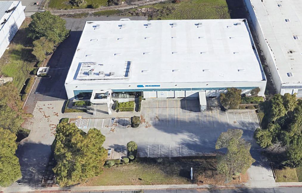 Top view of a large white warehouse that has a parking lot full of grass and trees around it.