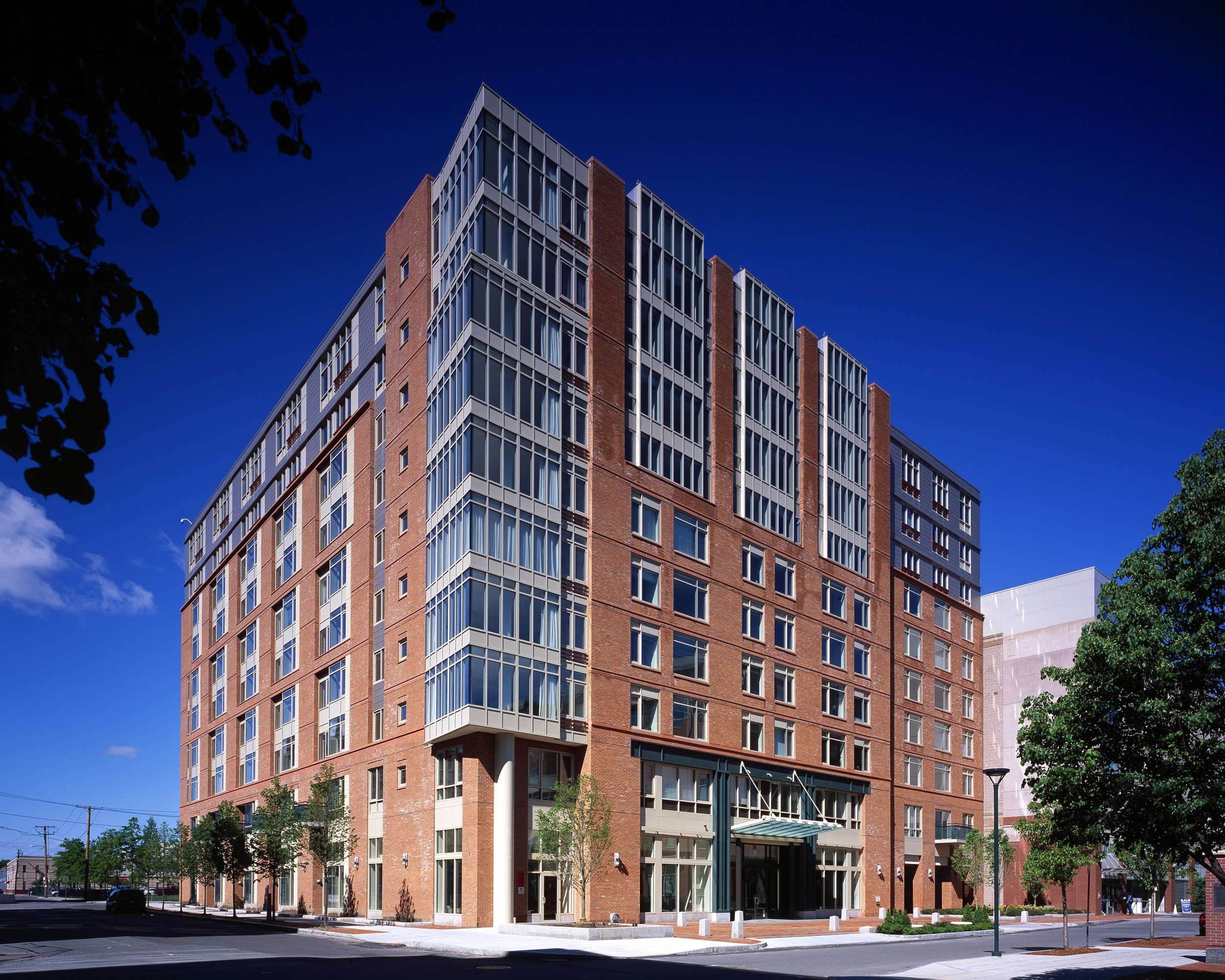 Tall apartment building in the middle of the city with trees surrounding the outdoor area.