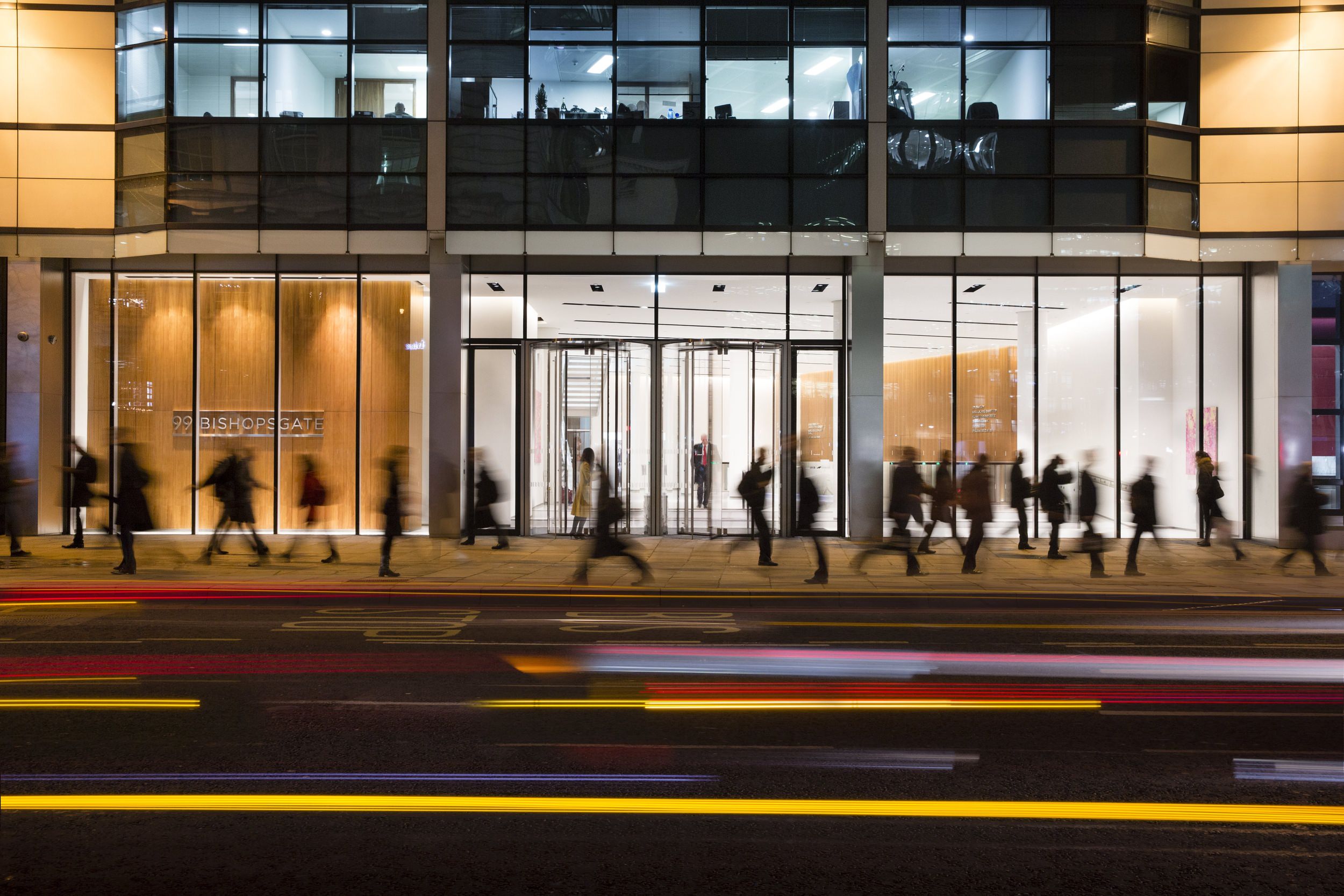 A crowd of people walking along the sidewalk that is in front of a building with glass panels.