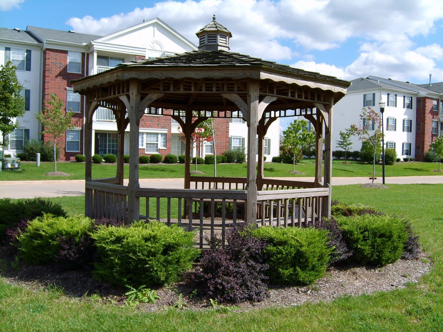 A gazebo in the middle of a park that is full of plants and trees with building in the background.