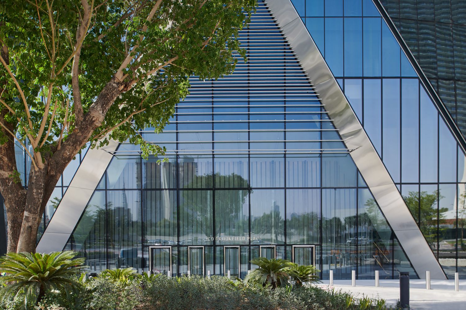 A large glass building that has an A shaped metal frame at the front of it and some trees out front as well.