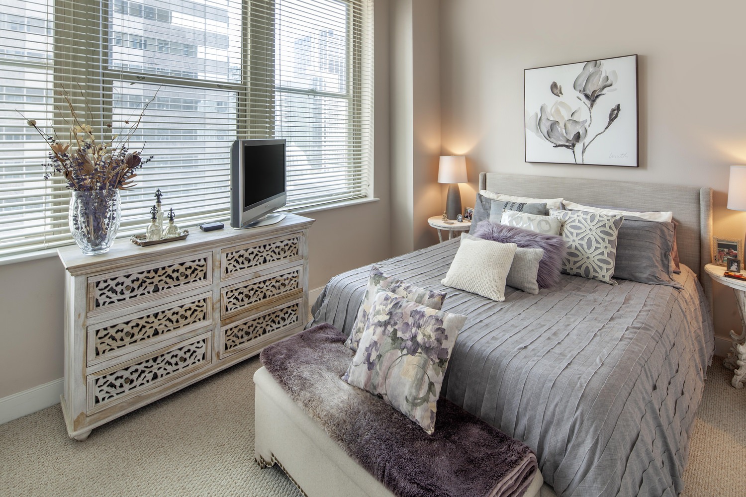 A bedroom that has a bed covered in grey sheets with a window showing the city outside.