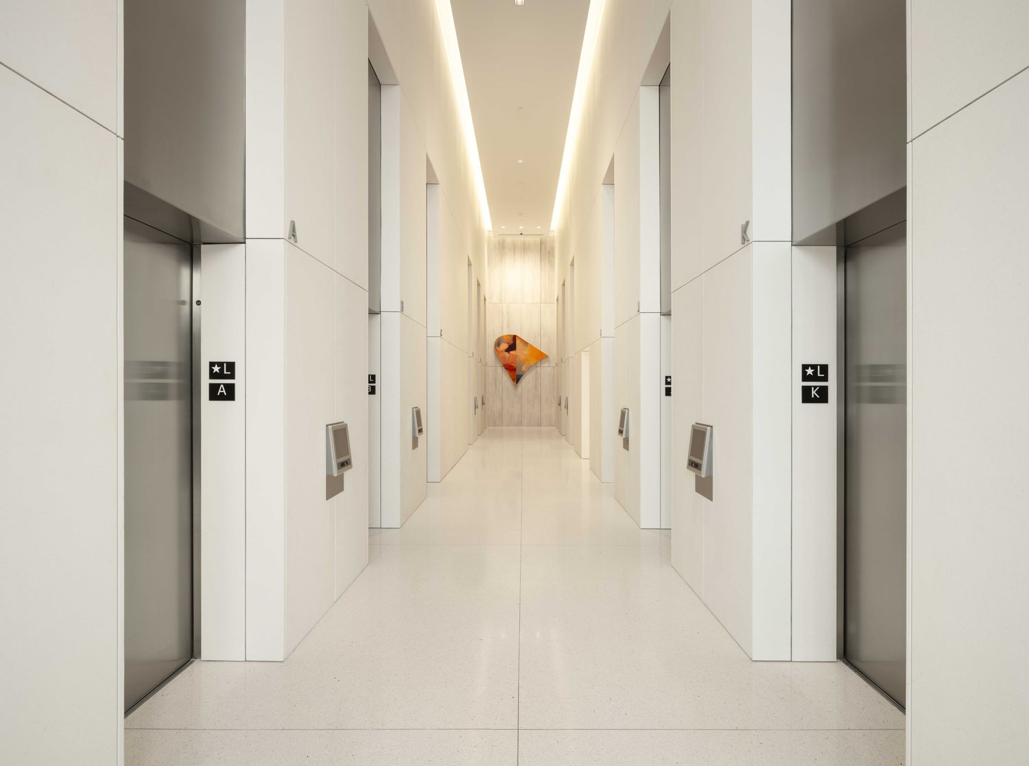 A hallway in an office building that has some elevators on either side of it and some white walls.