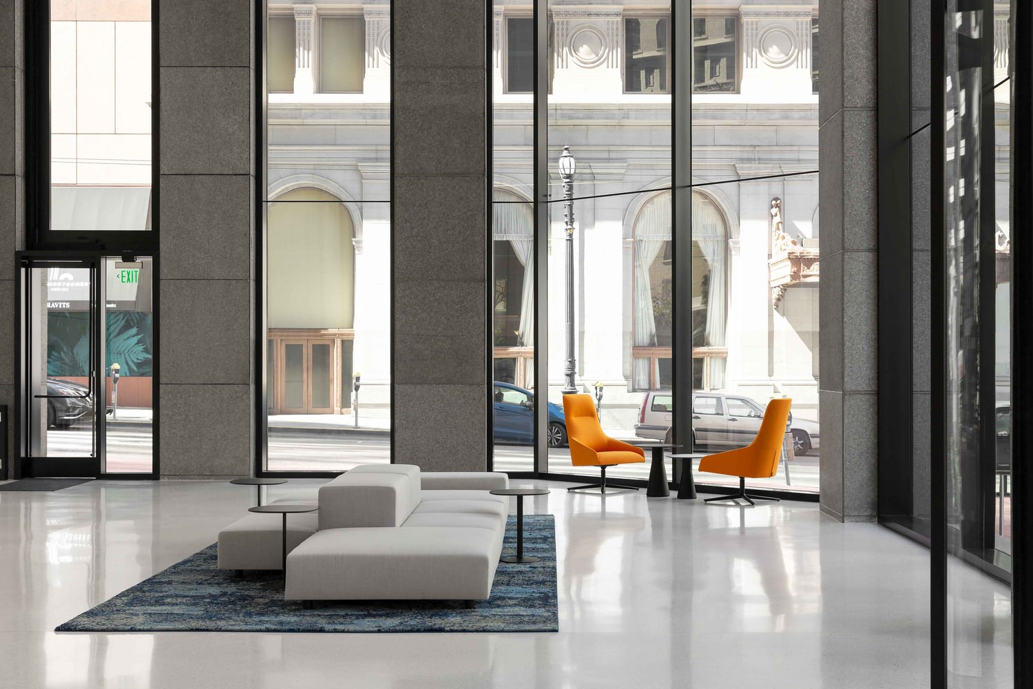 A lobby in a building that has a white couch and a table that has two orange chairs at it.