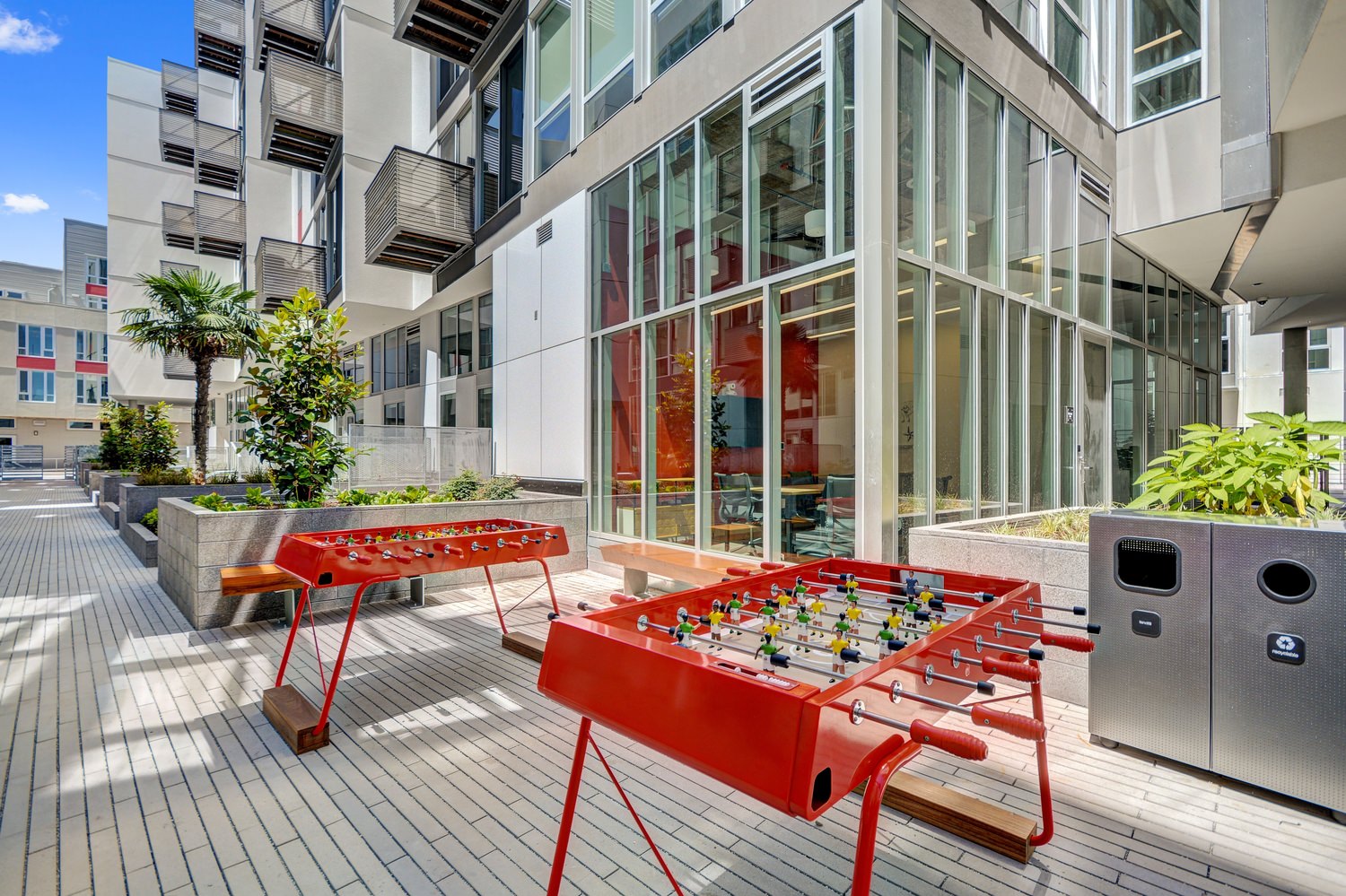 A walkway that is line with game tables and plants that is next to a large building with windows.