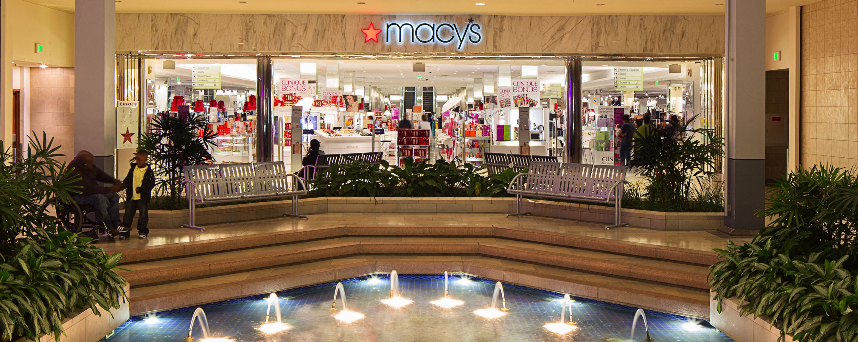 Fountains shoot out water in a fountain with benches in front of a Macy's located in a shopping mall.