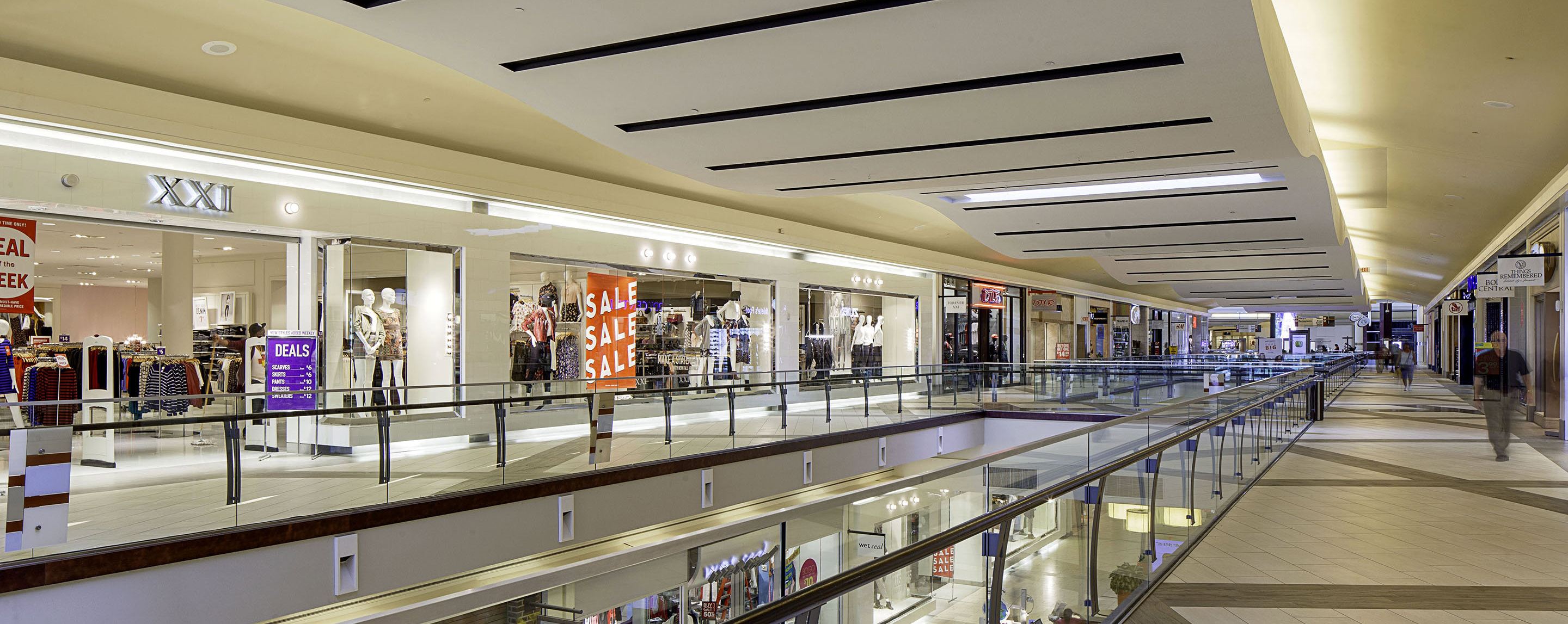Clothing stores line a long corridor in a brightly lit indoor multilevel shopping center.