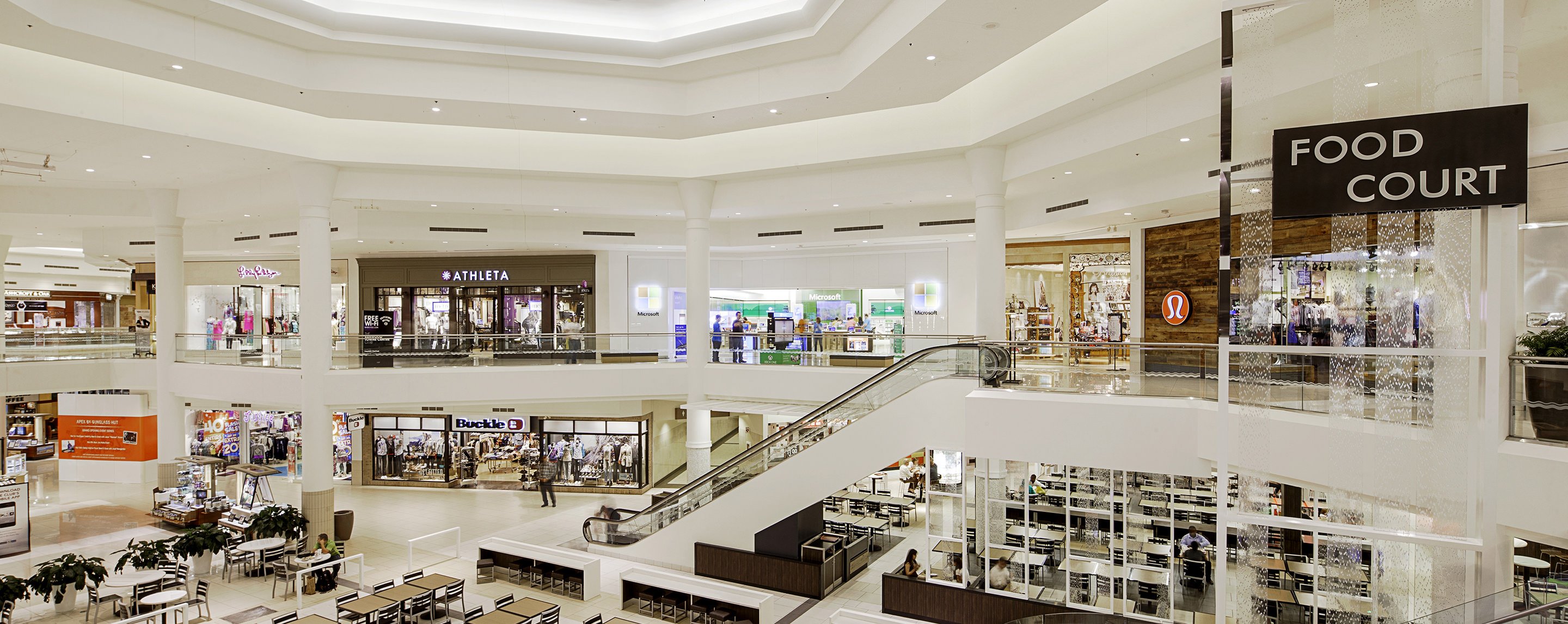 A wide view of an inner area of a two-story mall with Athleta, Lululemon, Buckle, and a Microsoft Store near an escalator.