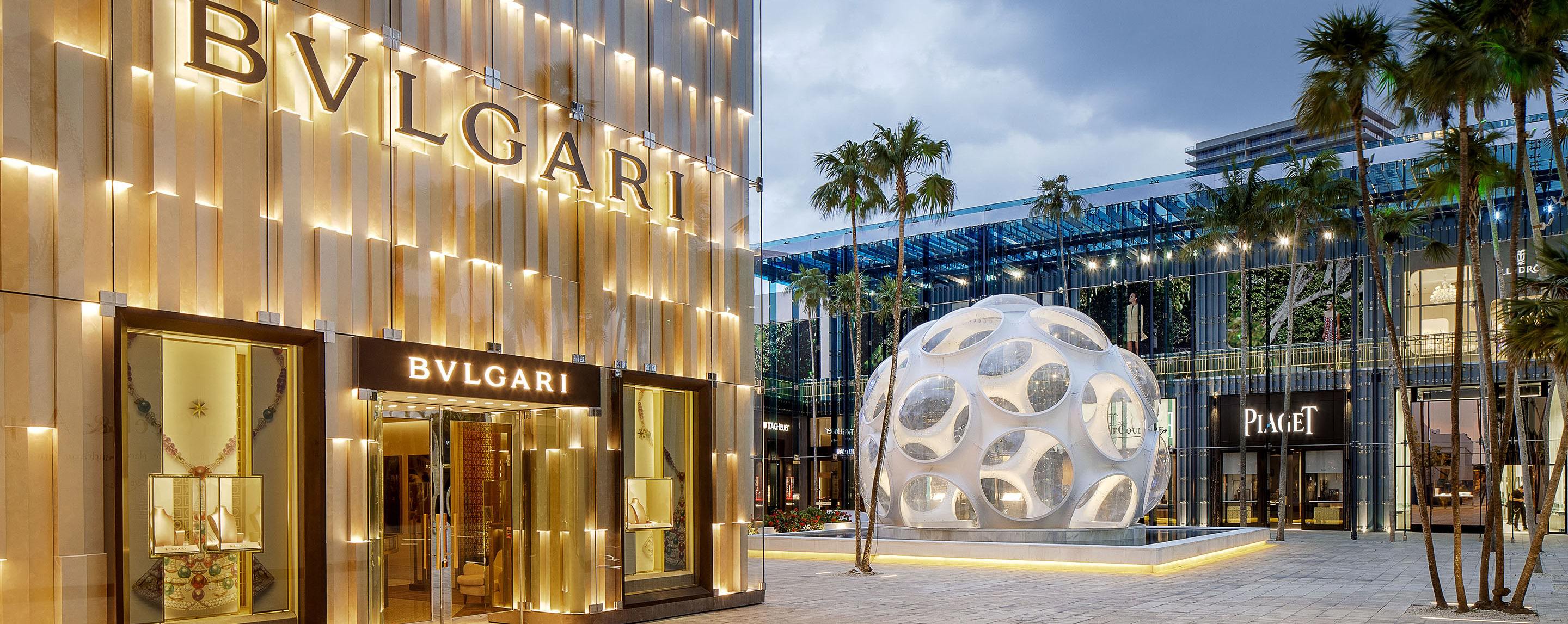 A large white sculpture stands to the side of a well lit Bvlgari storefront at twilight.