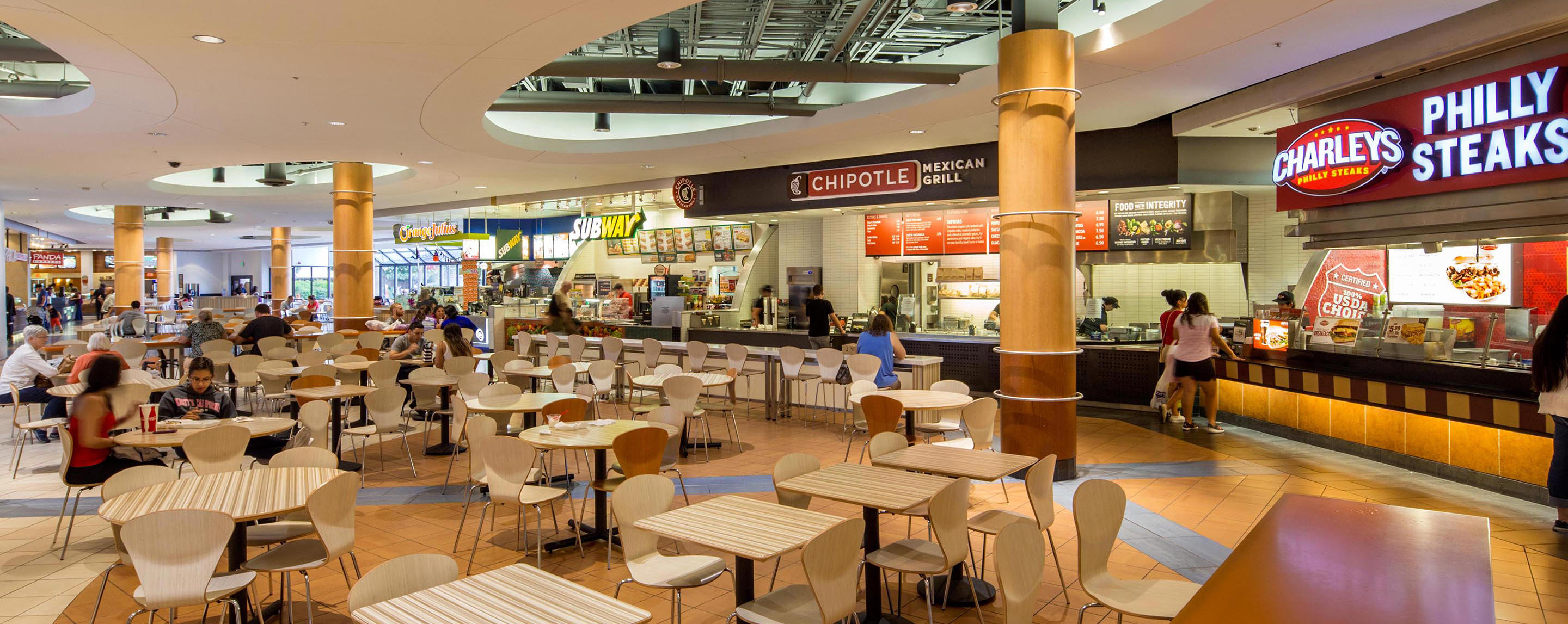 People sit at tables, walk around, and order food in a mall food court with establishments such as Subway and Chipotle.
