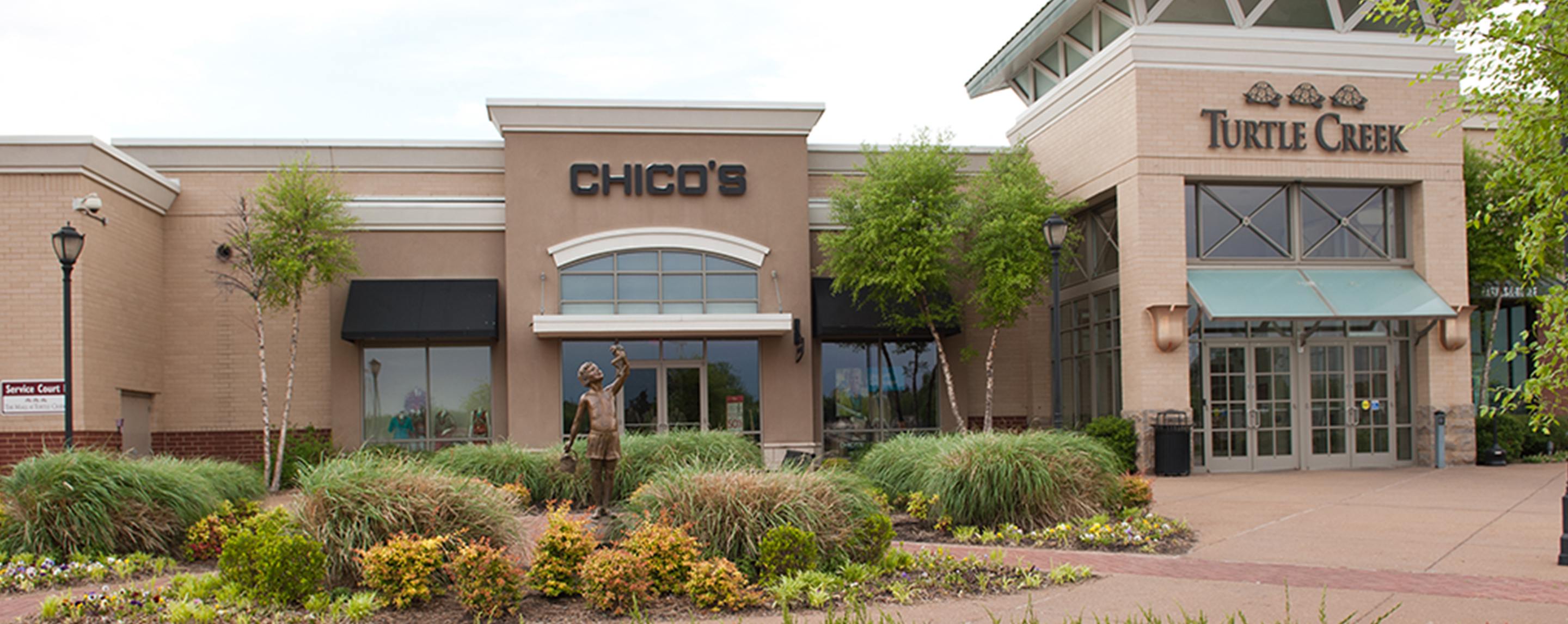 The exterior of a mall. There are bushes in the landscaping in front, and a sign for Chico's is visi