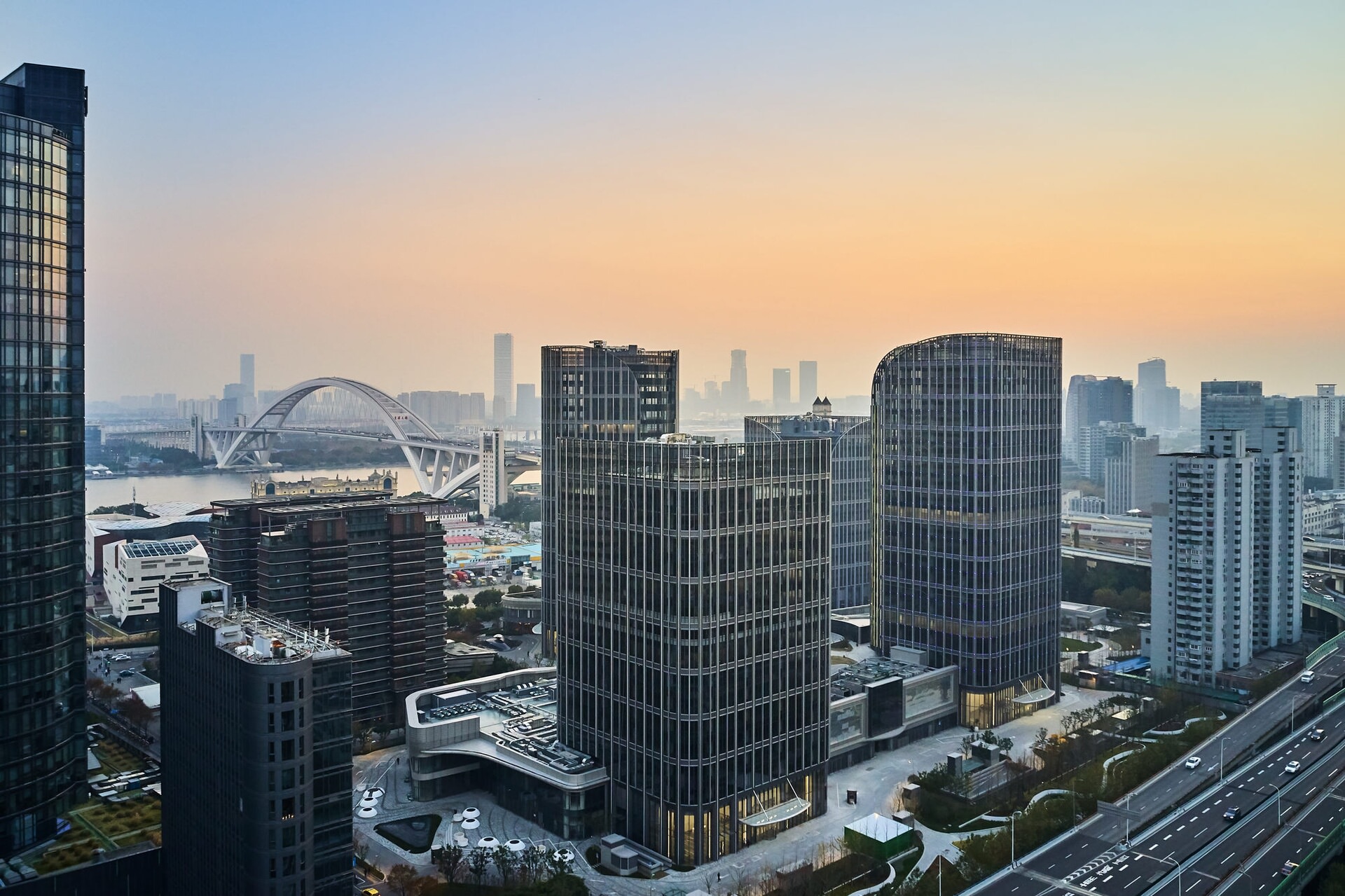 A row of tall buildings next to a busy city street that has a river and bridge in the background.