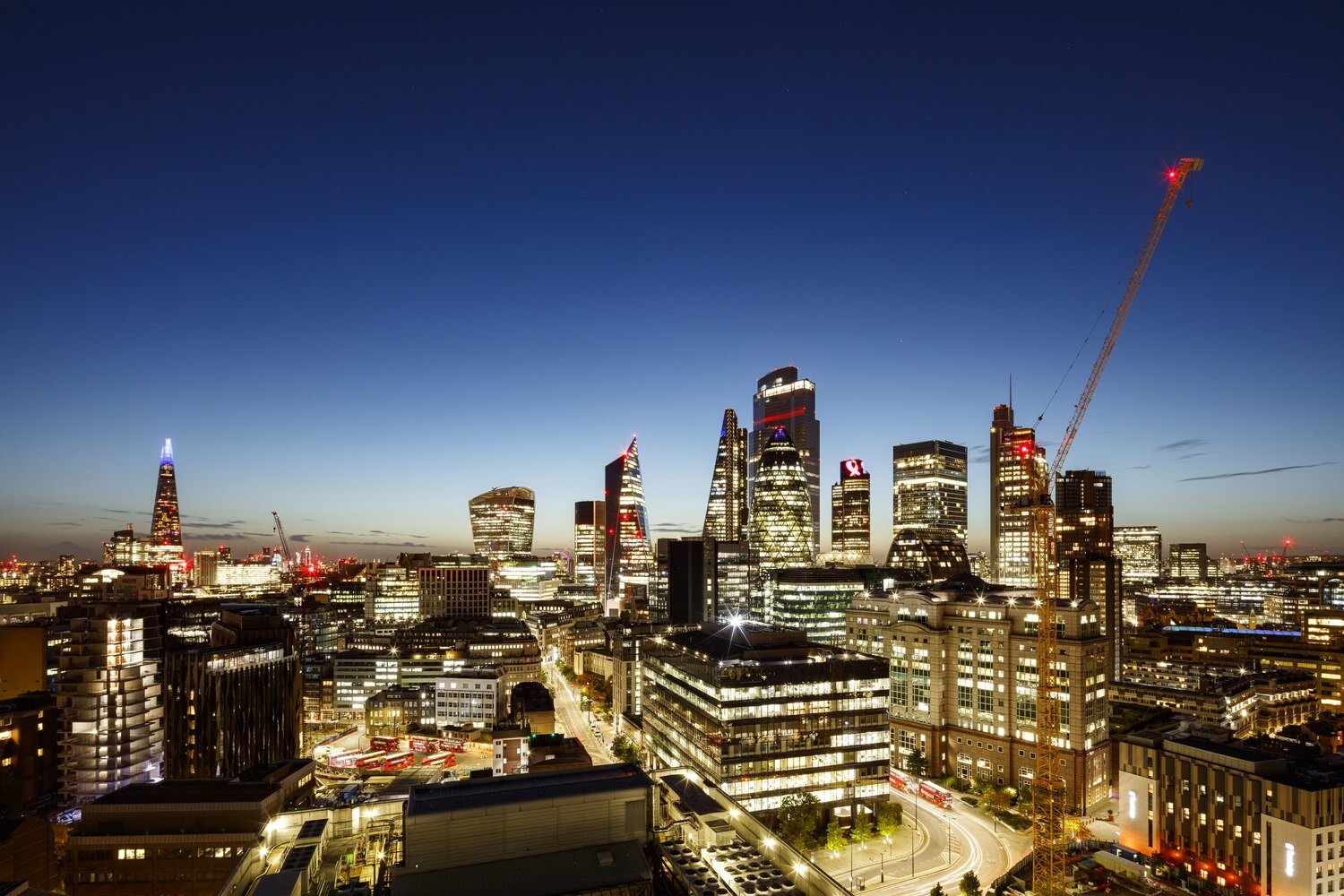 View of a busy city at night. All of the skyscrapers are lit bright, and there is a large crane in the foreground