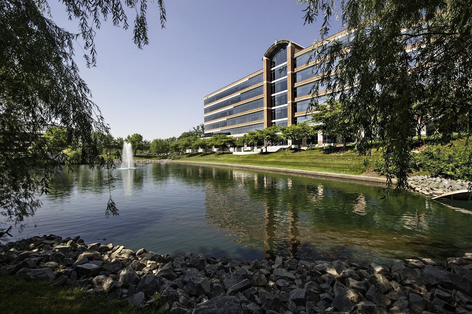 A big office building that is located next to a small lake that has a fountain in the middle of it.