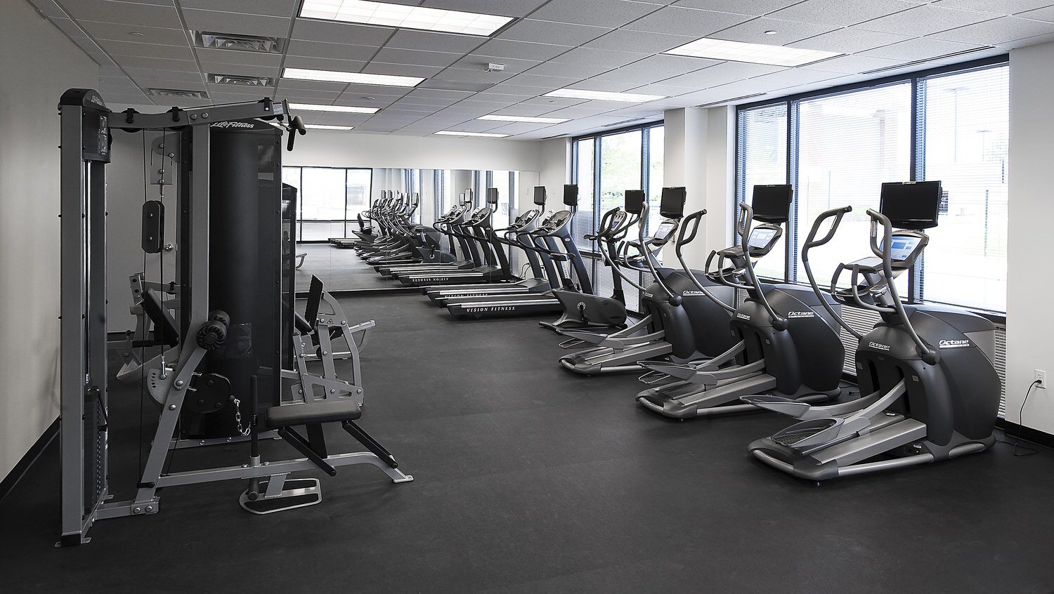 A lot of treadmills and elliptical machines that are located in a gym with rubber floors on it.