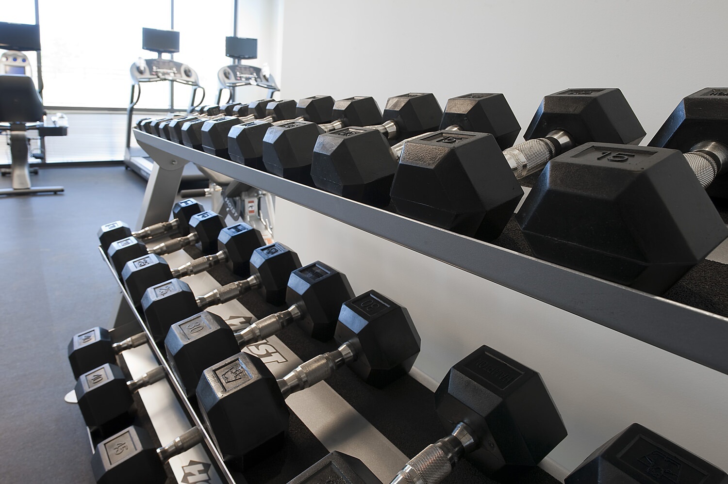 A gym that has barbells in a barbell rack and some treadmills and exercise machines behind them.