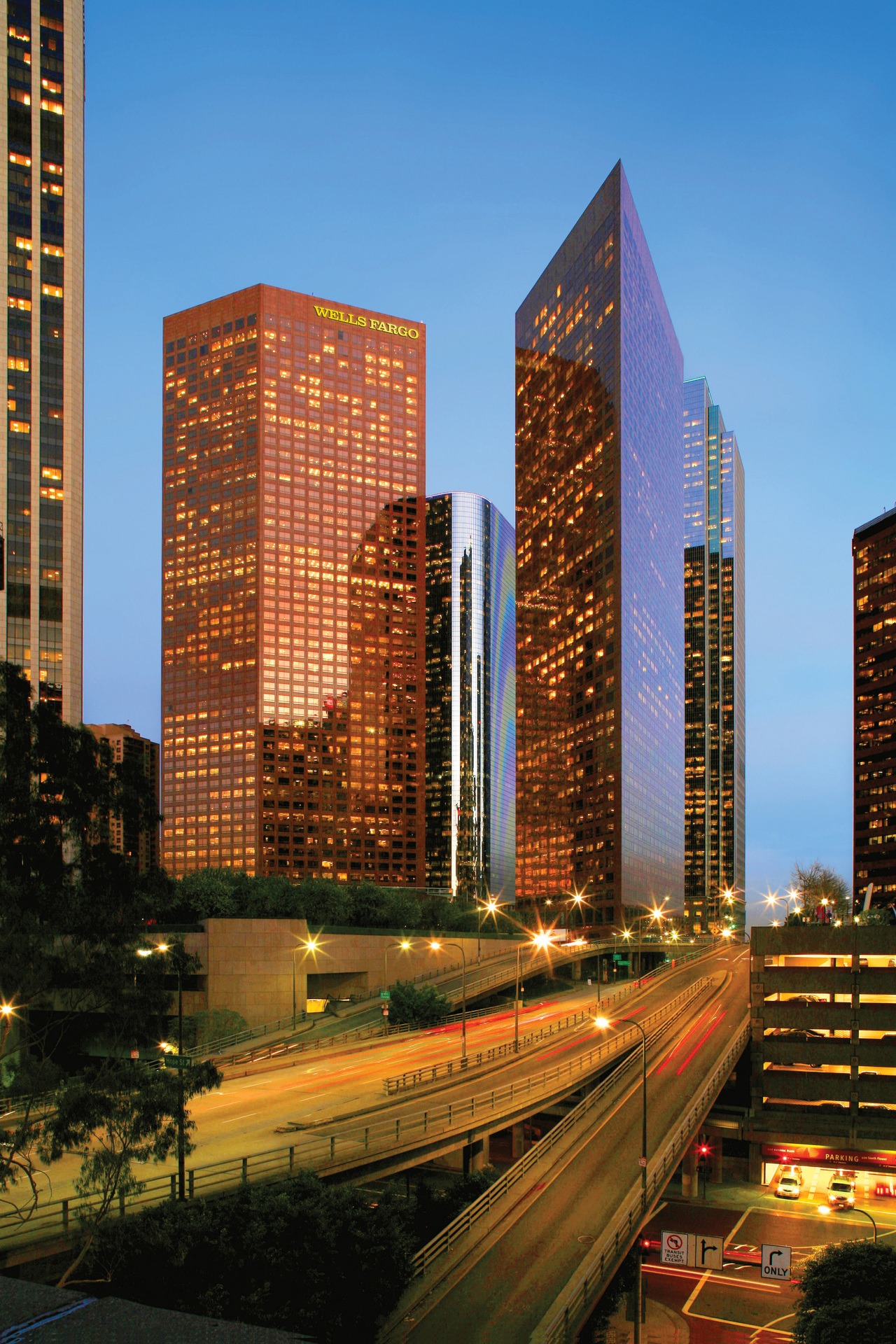 A busy city street with two red skyscrapers in the background at twilight. One of the buildings says "WELLS FARGO"